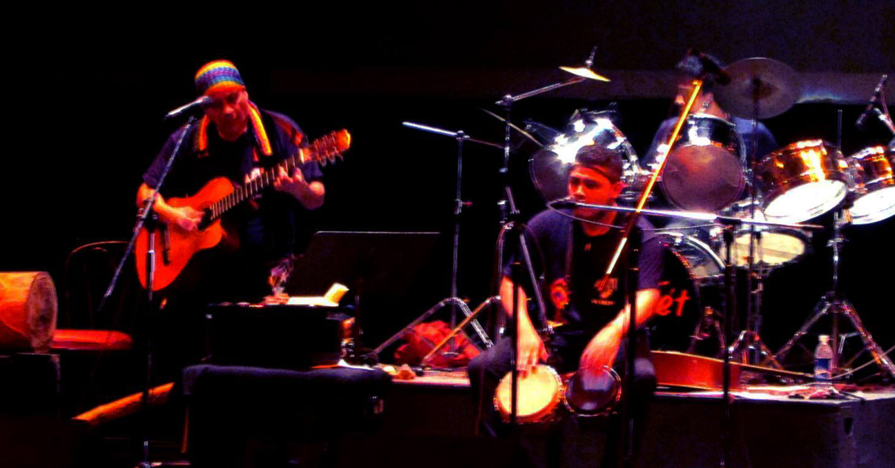 Tres hombres en un escenario. De izquierda a derecha, uno toca la guitarra, otro dos pequeños tambores y el último al fondo la batería.