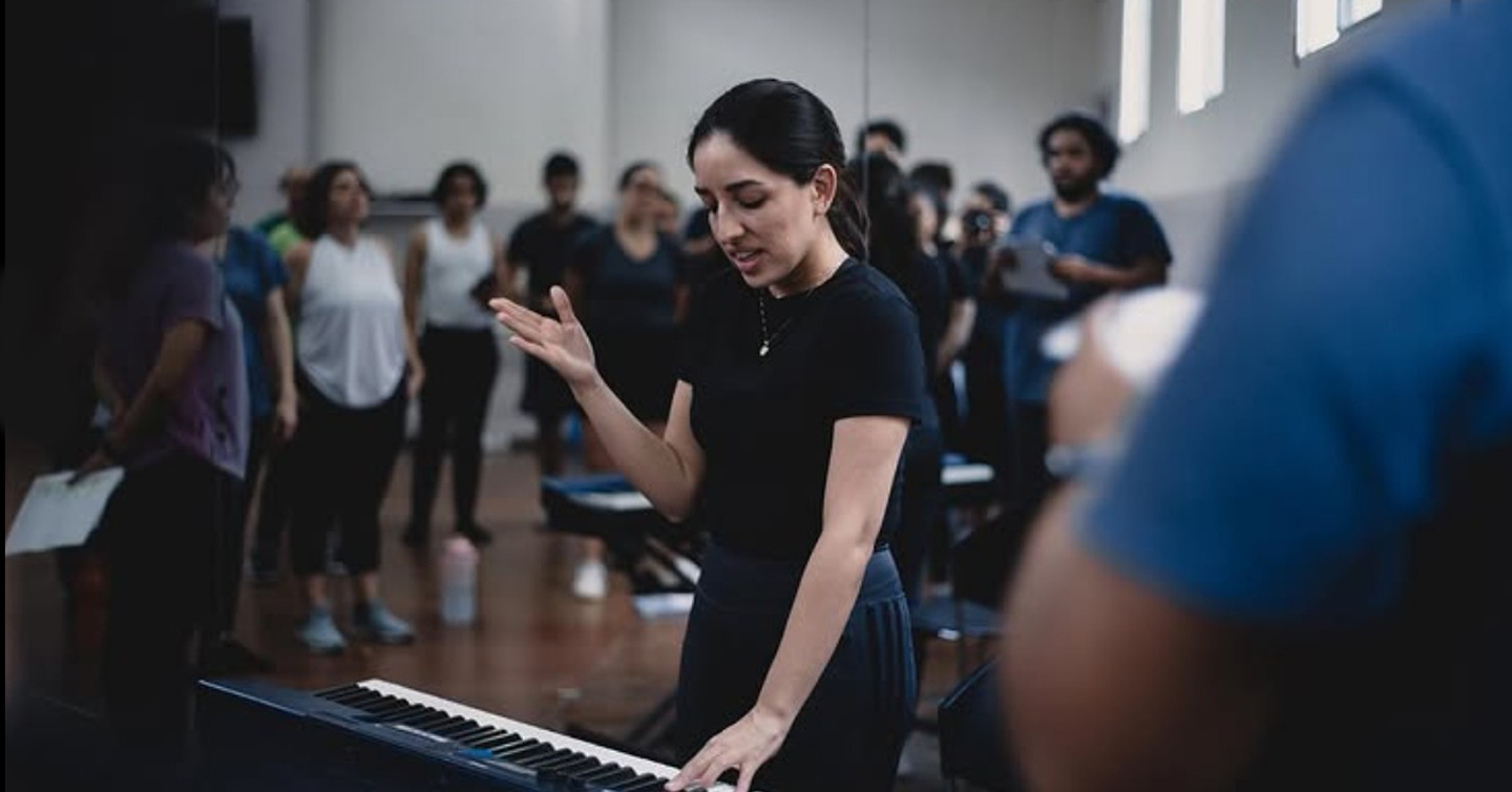 Maestra de canto toca notas en un teclado rodeada de alumanas y alumnos. 