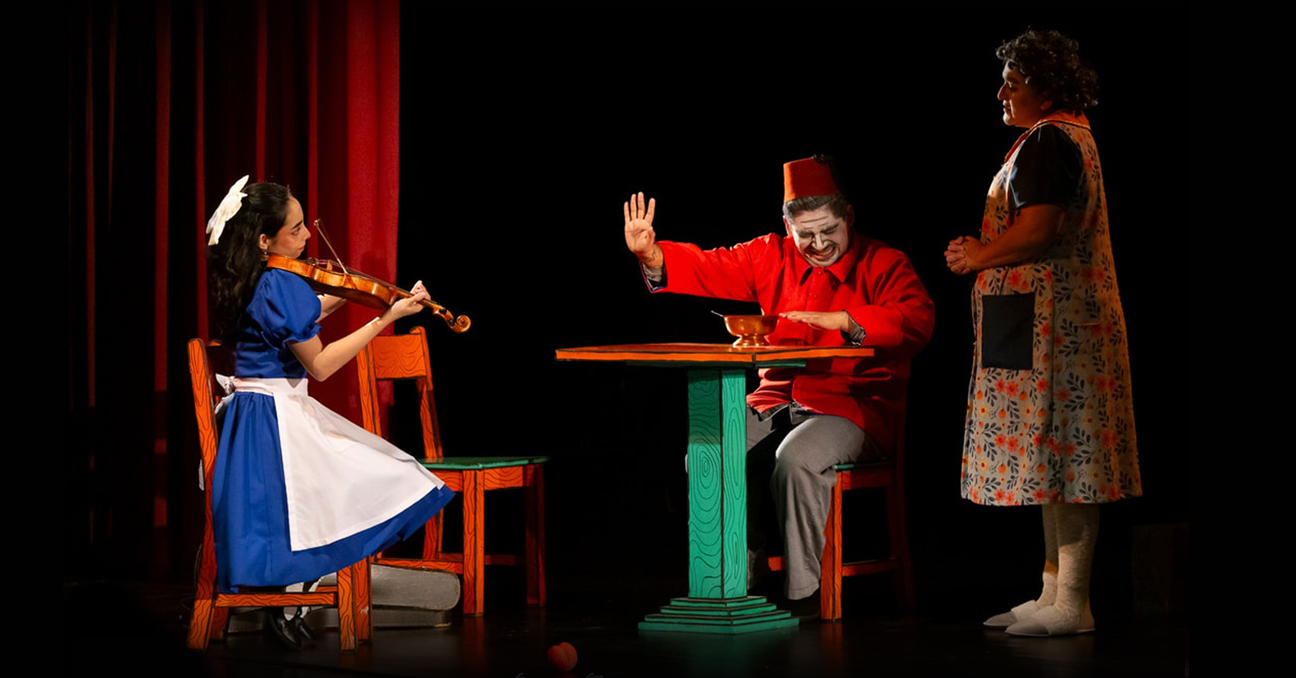 Tres actores de la obra "La metamorfosis" en el escenario. A la derecha, una mujer de mediana edad. En el centro, un hombre vestido de rojo. Y a la izquierda, una actriz simula ser una niña tocando el violín.