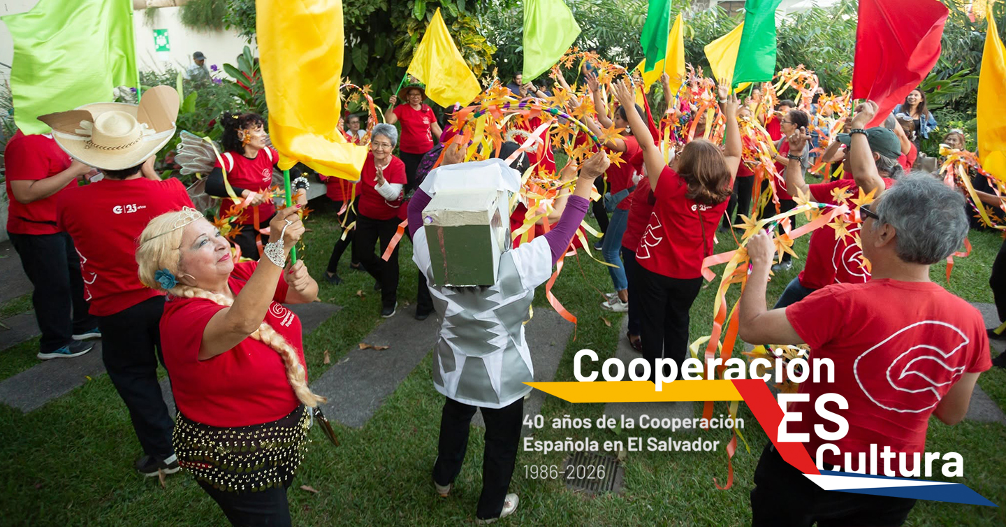 Un grupo grande de actrices y actores adultos mayores ondean banderas de colores. El texto dice: Cooperación ES Cultura, 40 años de la Cooperación Española en El Salvador (1986-2026)
