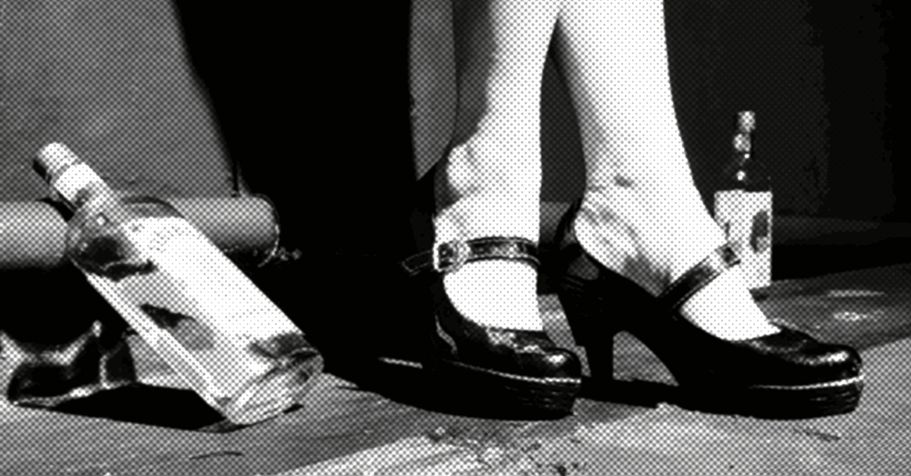 Una fotografía en blanco y negro con los pies de una mujer en unos zapatos de tacón, como de baile, y una botella de licor en el piso